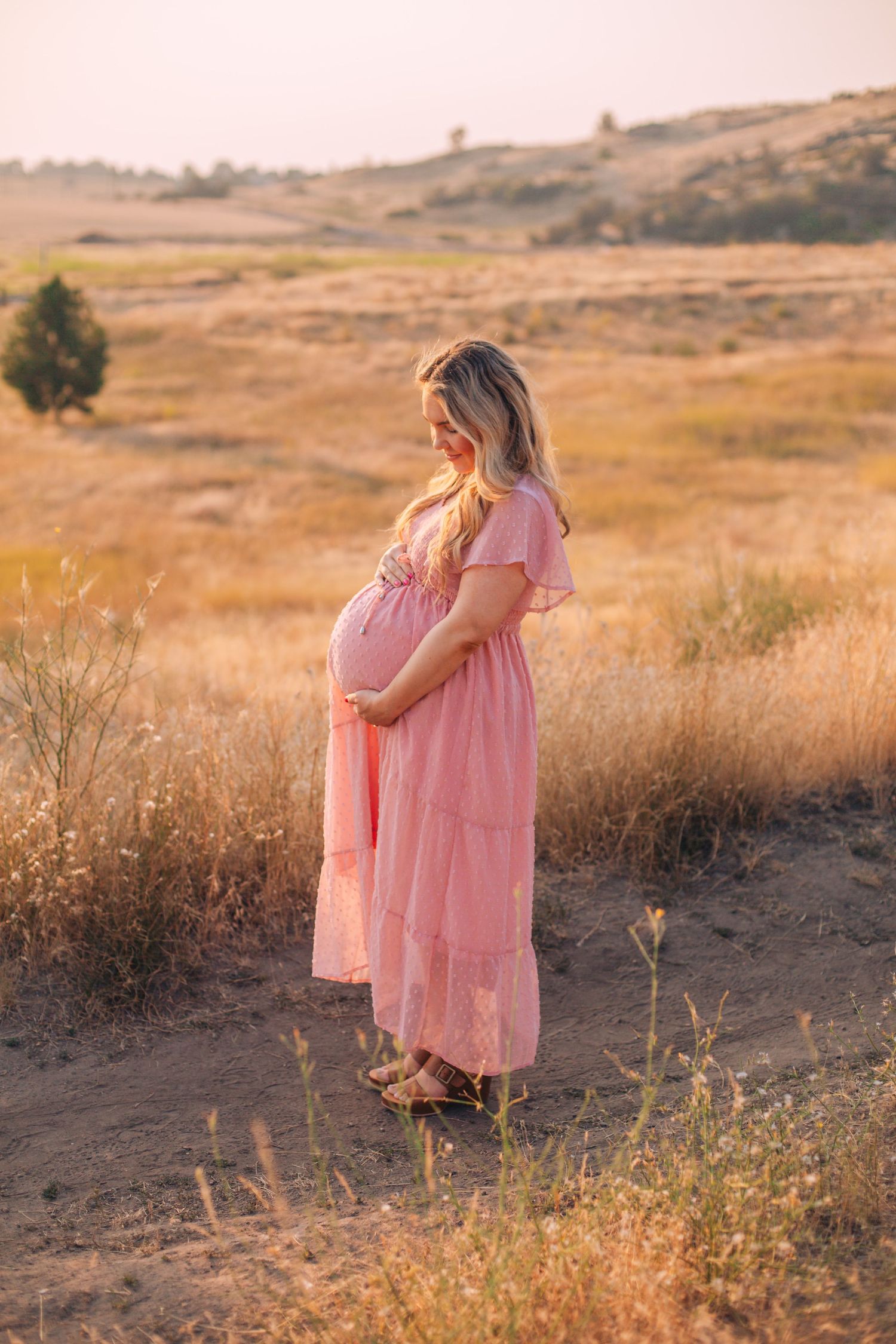 Pregnant woman in pink dress holding belly in golden field at sunset.