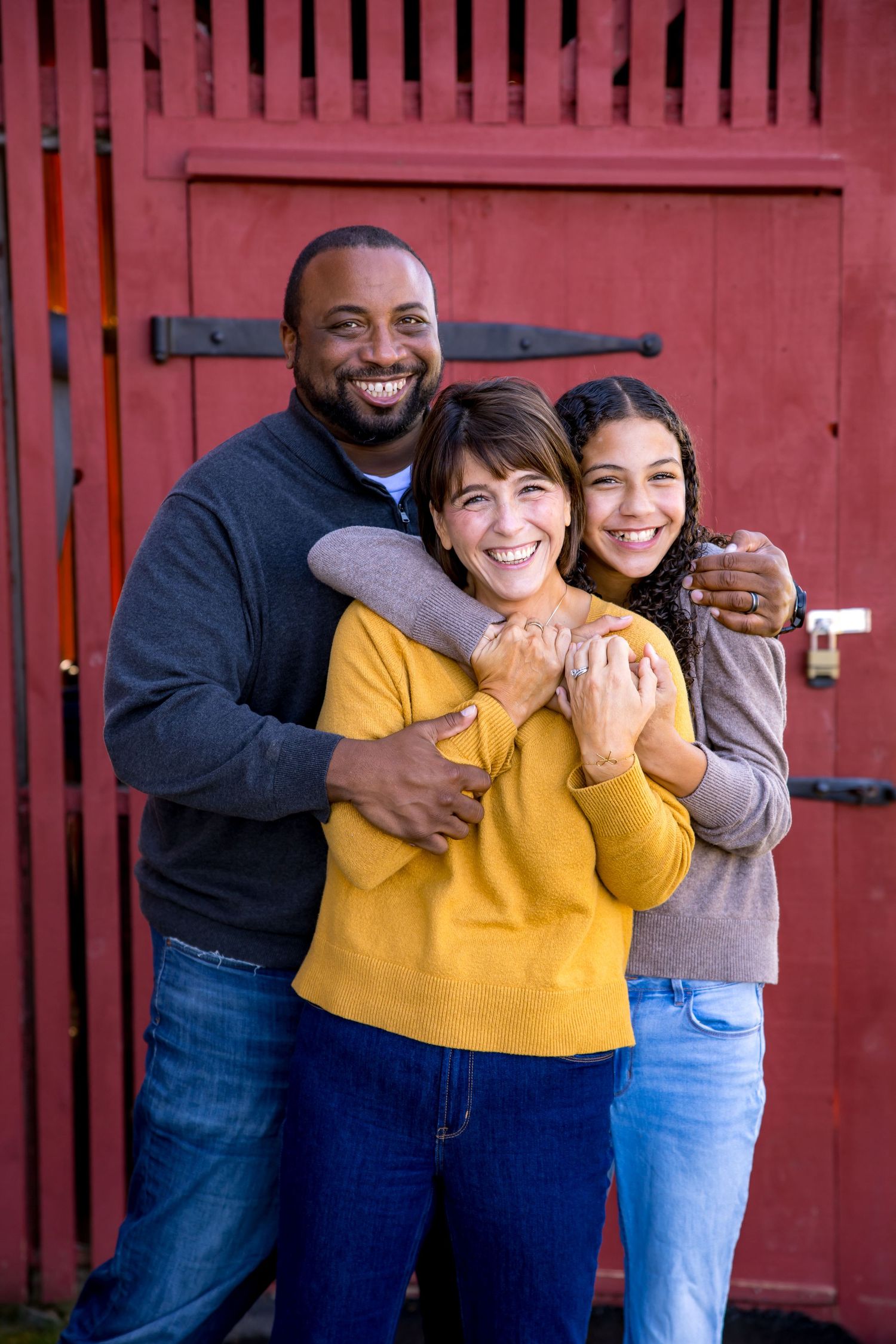 A smiling multiracial family of three hugging warmly in front of a red barn door.