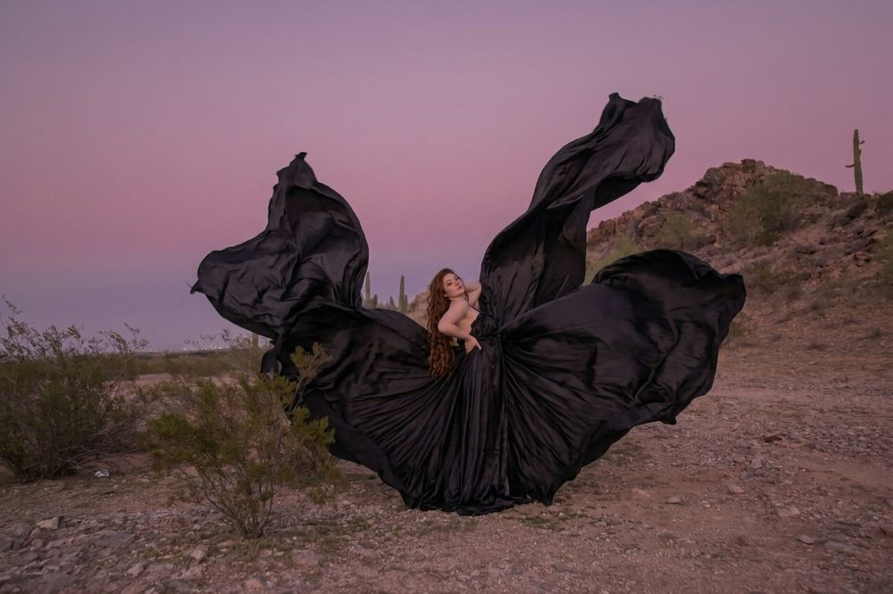 Woman in dramatic black flowing gown poses in desert landscape at sunset with fabric billowing in wind.