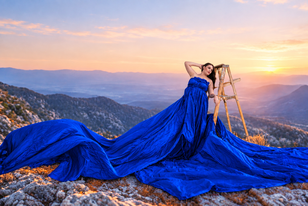 Woman in flowing royal blue gown poses on rocky mountain summit at golden sunset with panoramic valley views.