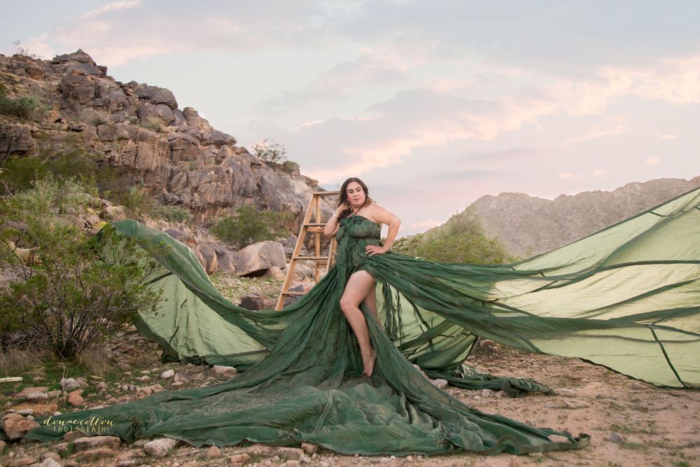 Woman in flowing green gown poses dramatically in desert landscape with rocky mountains at sunset.