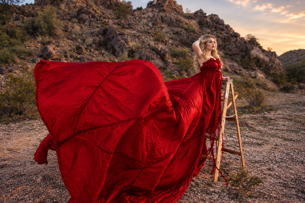A woman in a flowing red gown poses dramatically in a desert landscape with rocky hills at sunset.