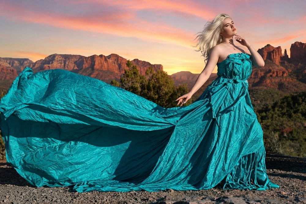 Blonde woman in flowing teal gown poses against stunning red rock canyon sunset landscape.