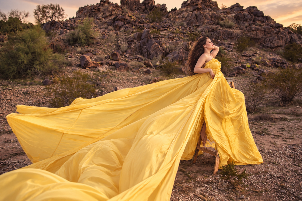 Woman in flowing yellow gown poses dramatically in desert landscape with rocky mountains backdrop.