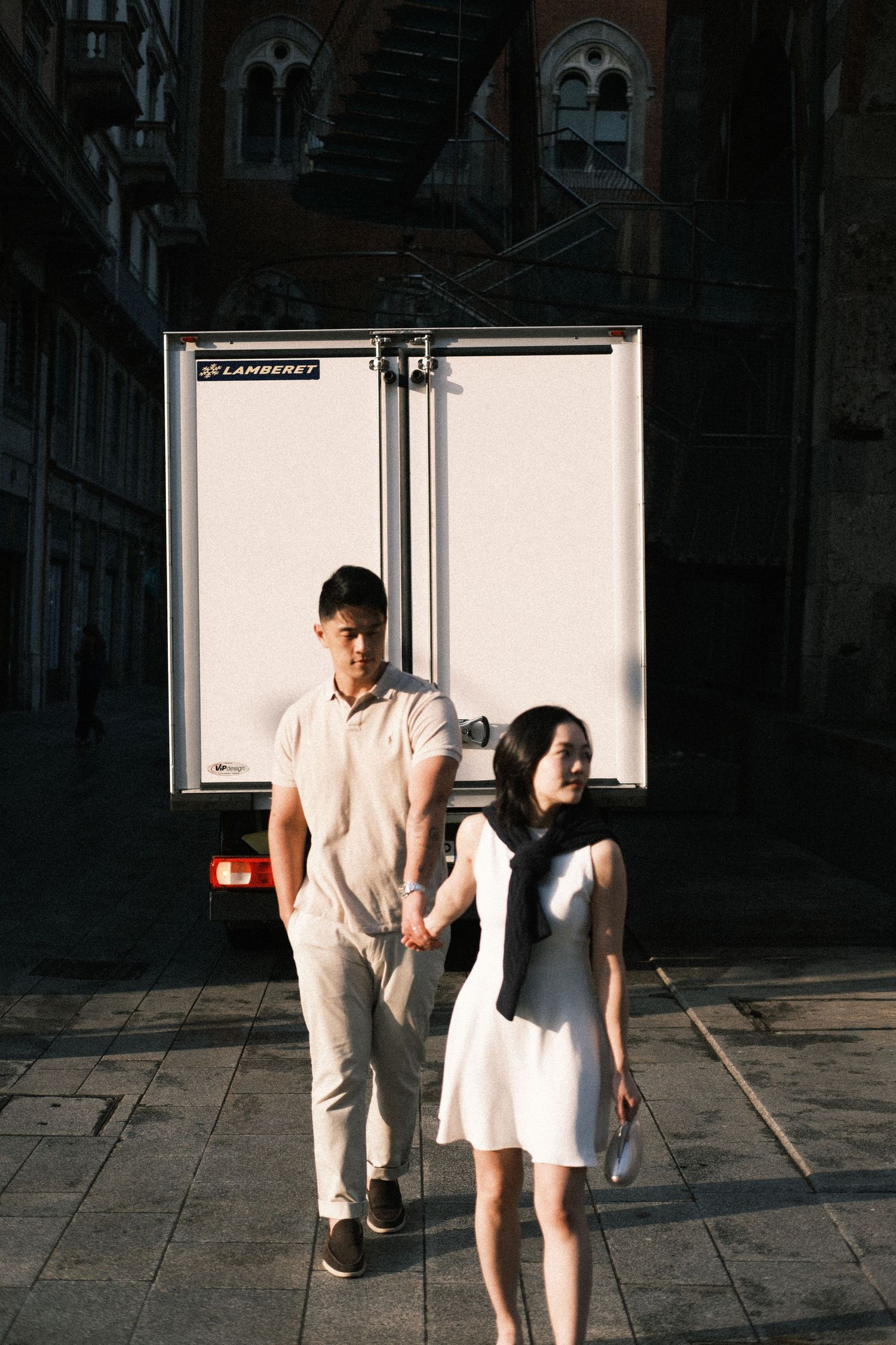 Couple holding hands walking in front of a white delivery truck on a sunlit cobblestone street.