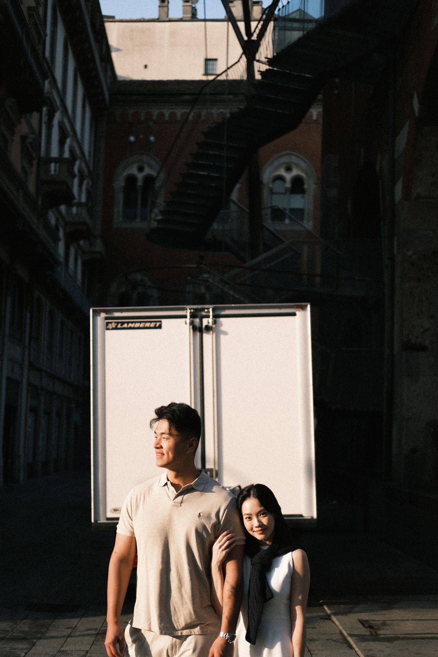 A young Asian couple poses together in an urban alleyway with a white truck and spiral staircase behind them.
