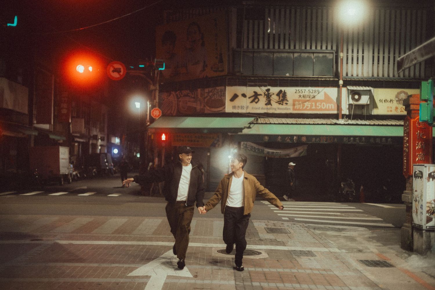 Two people crossing a street at night in Taiwan, with red traffic lights and Chinese signs illuminating the scene.