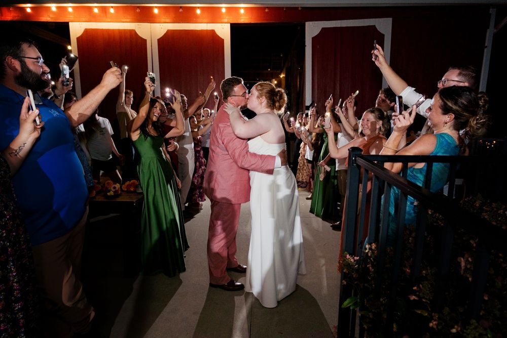 A bride in white and groom in a pink suit share a kiss during a cell-phone-lights non-sparkler send-off outside a Michigan barn venue.