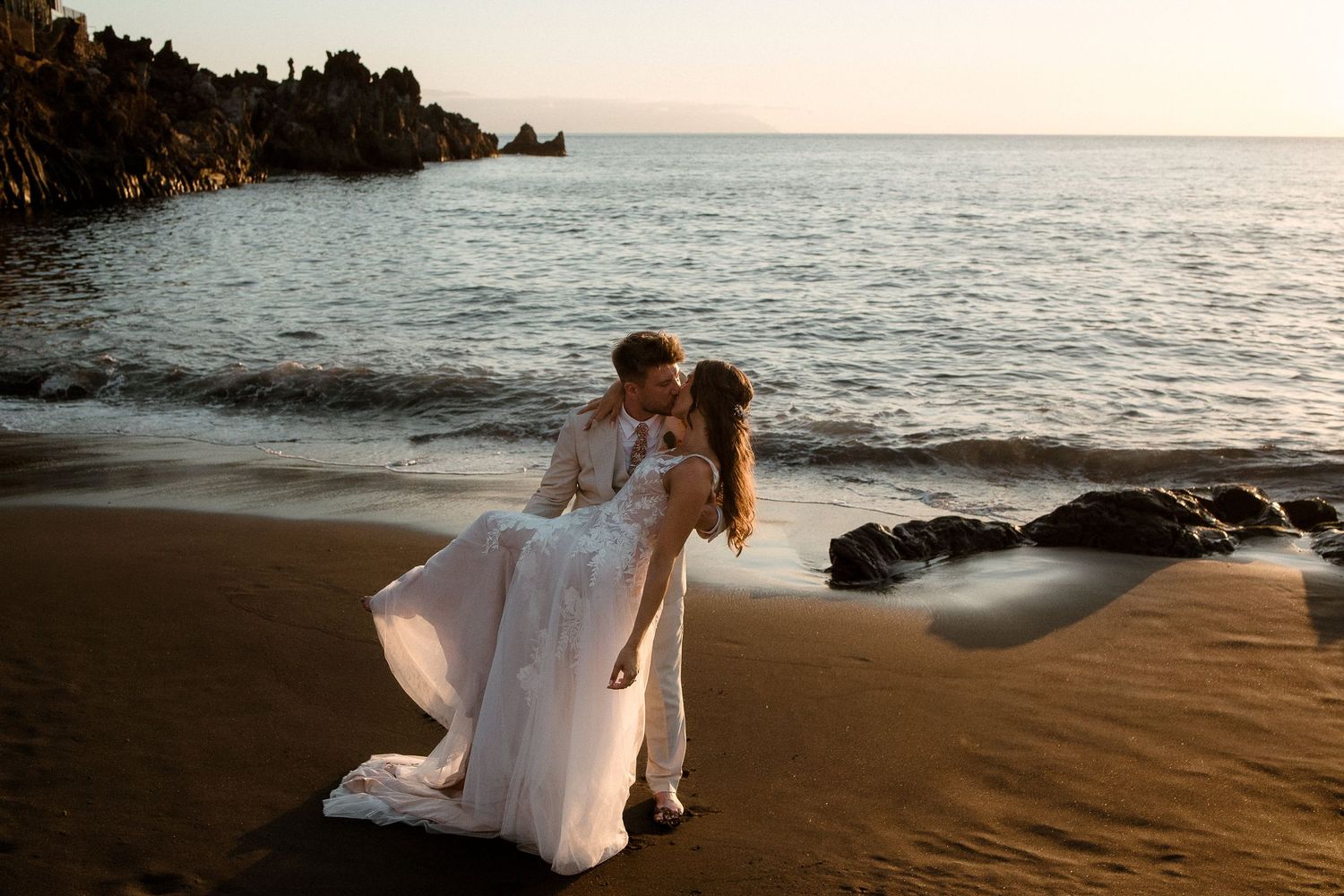 Bride and groom share a romantic kiss on a black sand beach at sunset with ocean waves in the background.