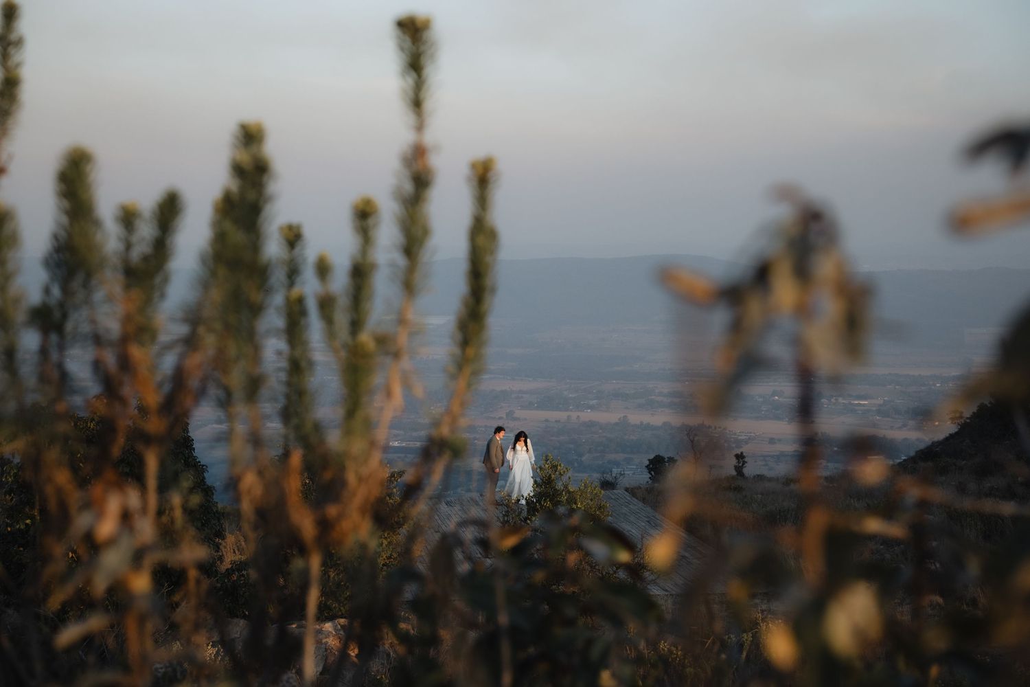 Couple in wedding attire standing on hilltop at golden hour sunset.