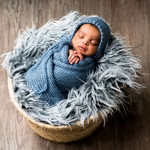 Newborn baby sleeping peacefully in a basket wrapped in a blue knit swaddle and bonnet on a fluffy gray fur blanket.