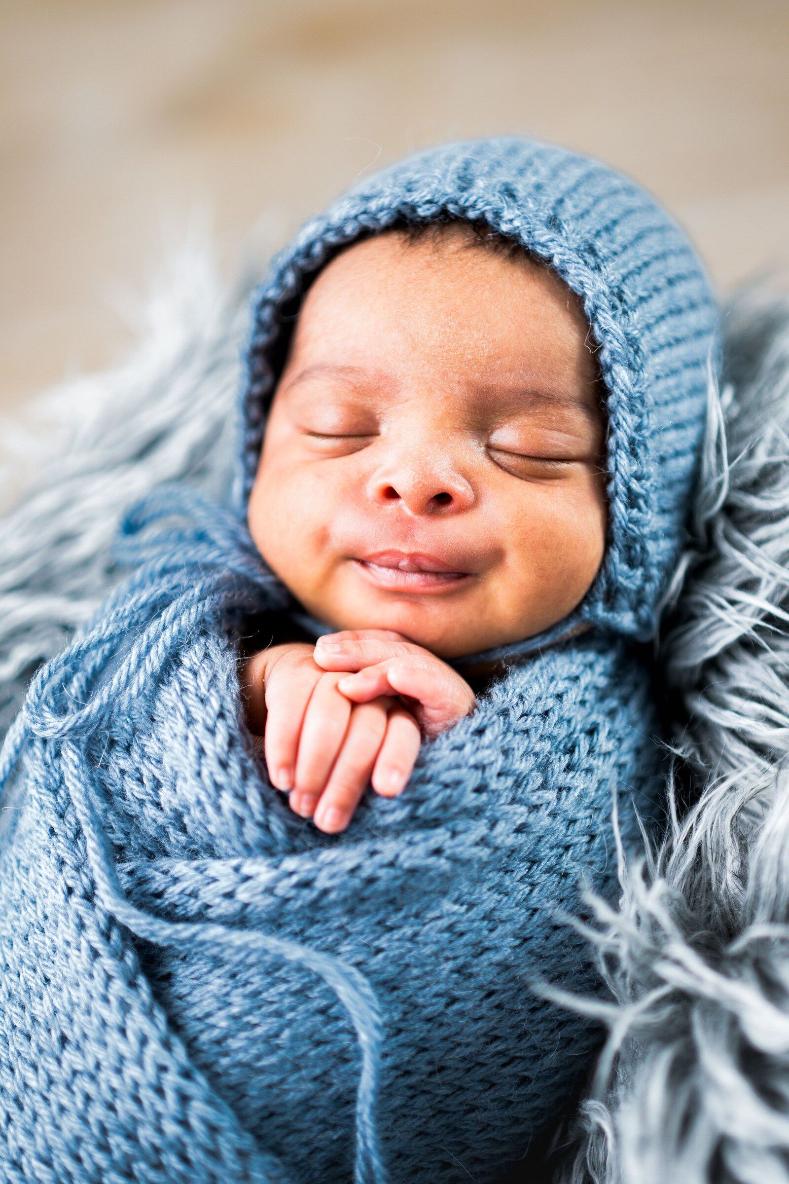 Sleeping newborn baby wrapped in a blue knit swaddle and matching bonnet, resting peacefully on a soft fur blanket.