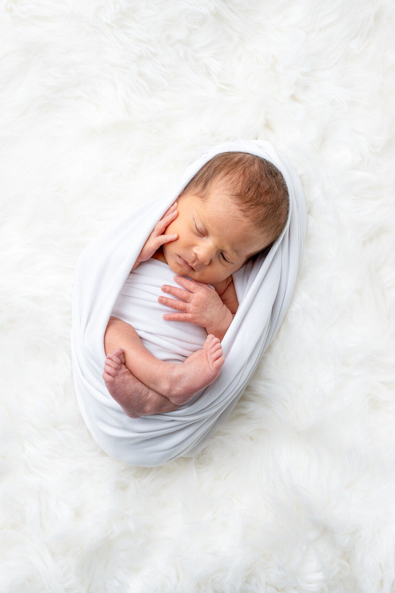 Newborn baby swaddled in white wrap sleeping peacefully on soft white feather blanket.