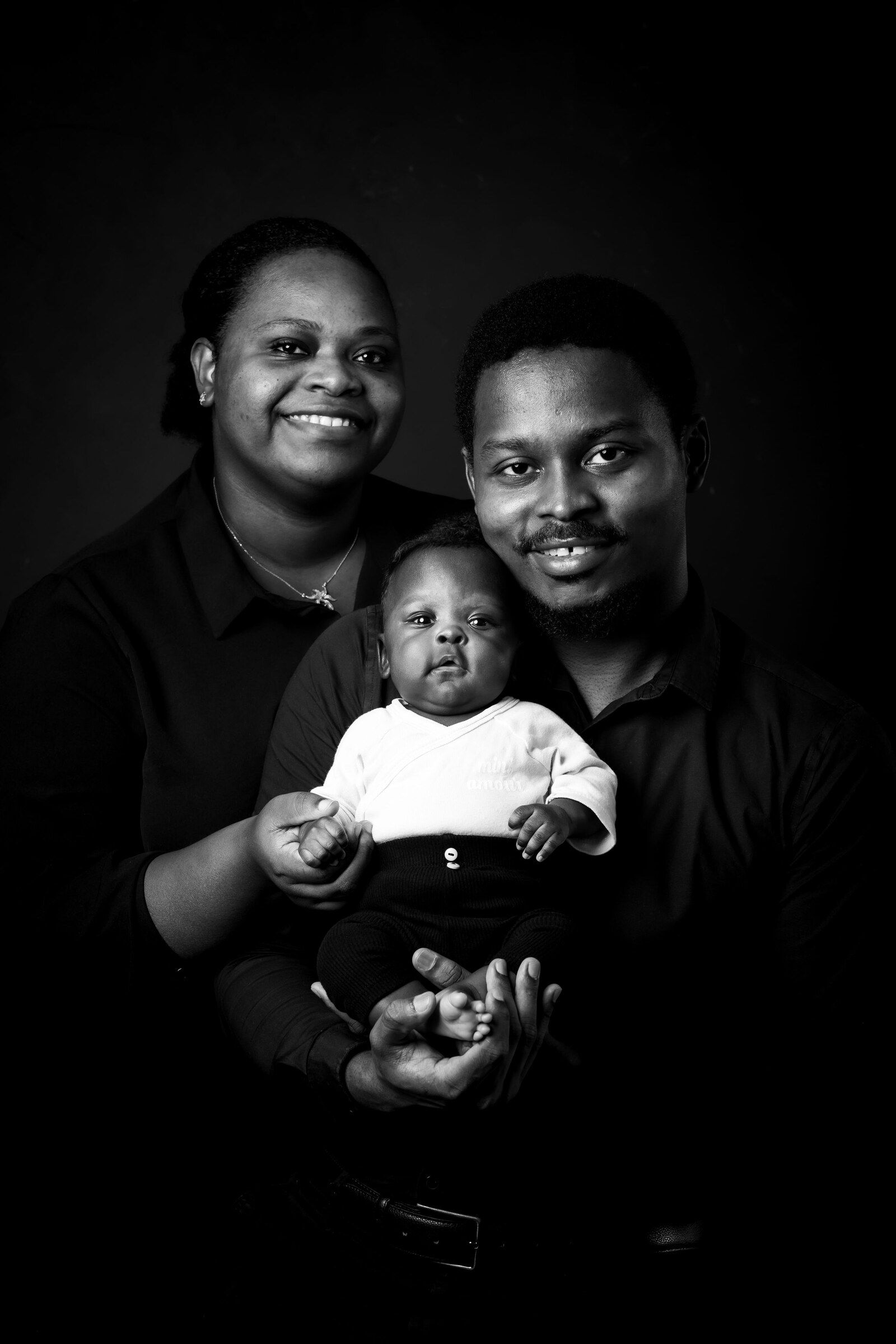 Black and white family portrait of smiling parents holding their infant baby against a dark studio background.