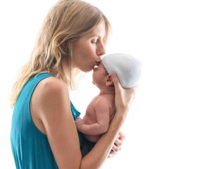 A mother in a teal dress kisses her newborn baby wearing a white hat against a white background.
