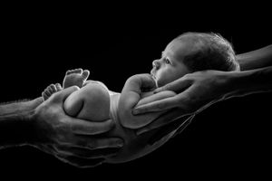 Black and white photo of a newborn baby cradled gently in two adult hands against a dark background.
