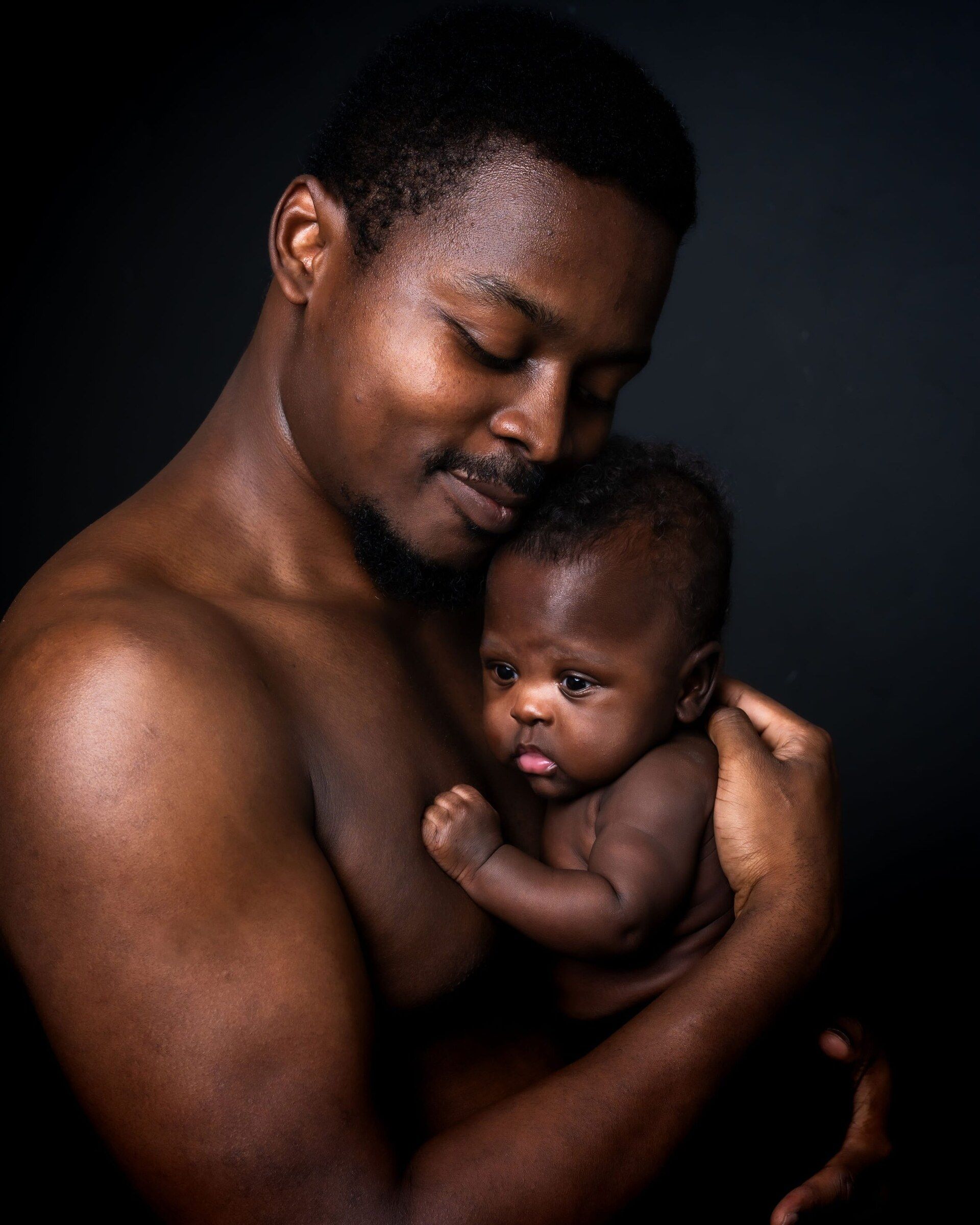 A shirtless Black father tenderly holds his newborn baby against his chest in a dark studio portrait.