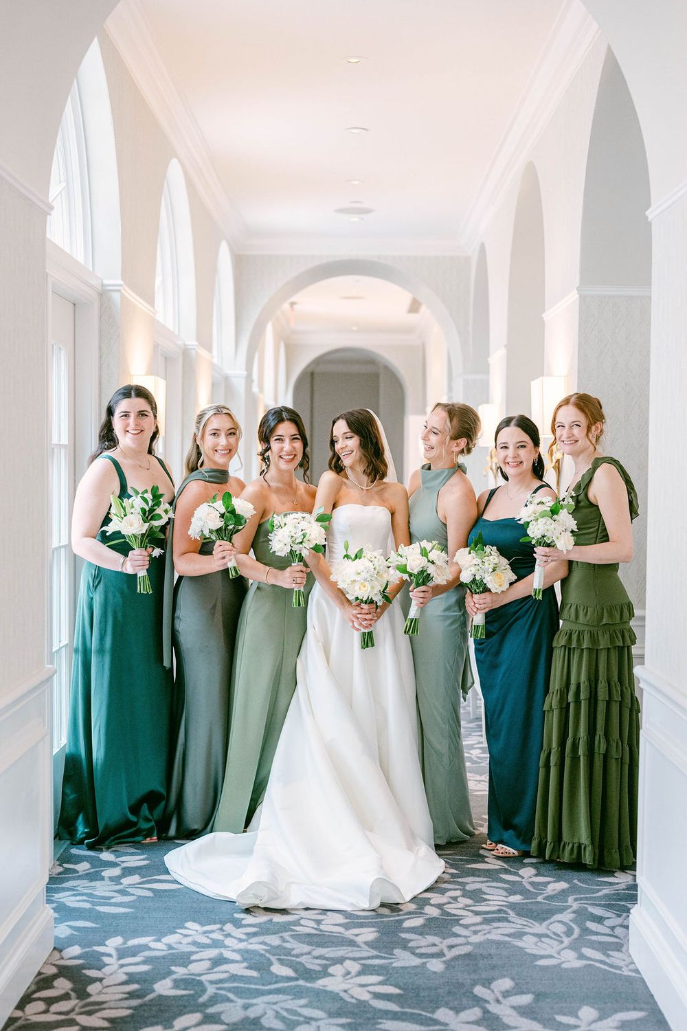 Bride in white gown poses with six bridesmaids in mismatched green dresses holding white bouquets in elegant hallway.