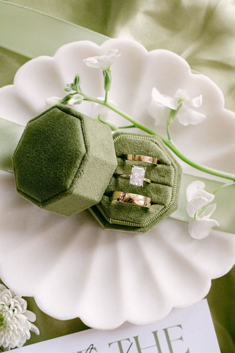 Green velvet hexagon ring box with gold wedding bands and diamond engagement ring on white floral plate.