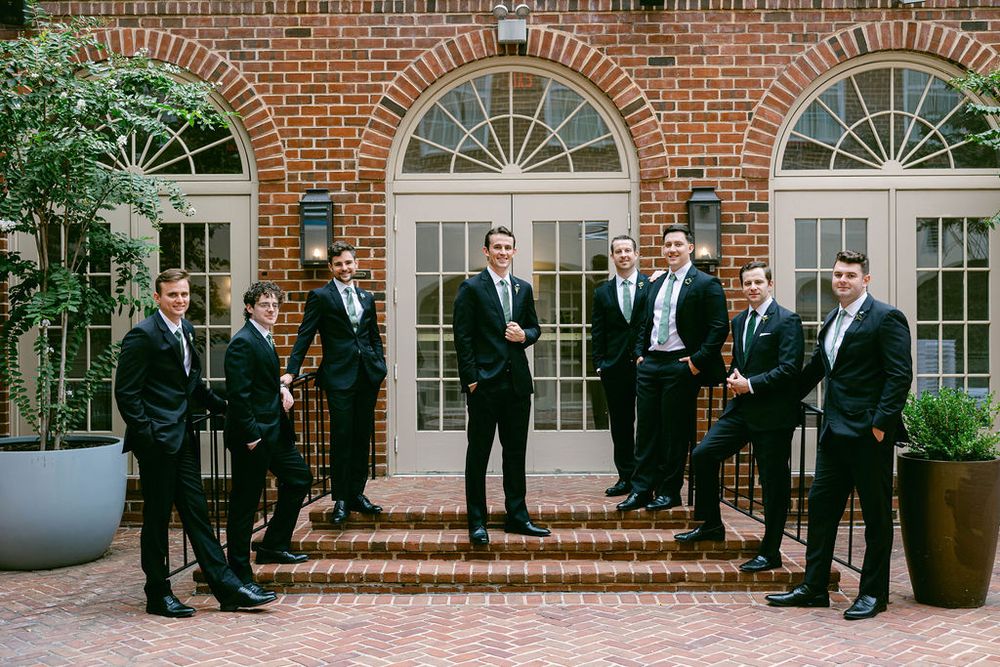 Groomsmen in black suits pose on brick steps outside an elegant brick building with arched windows.