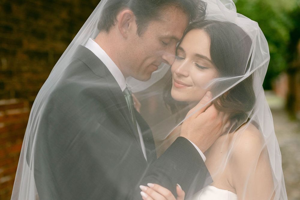 Bride and groom share an intimate moment under a flowing white wedding veil, embracing tenderly outdoors.