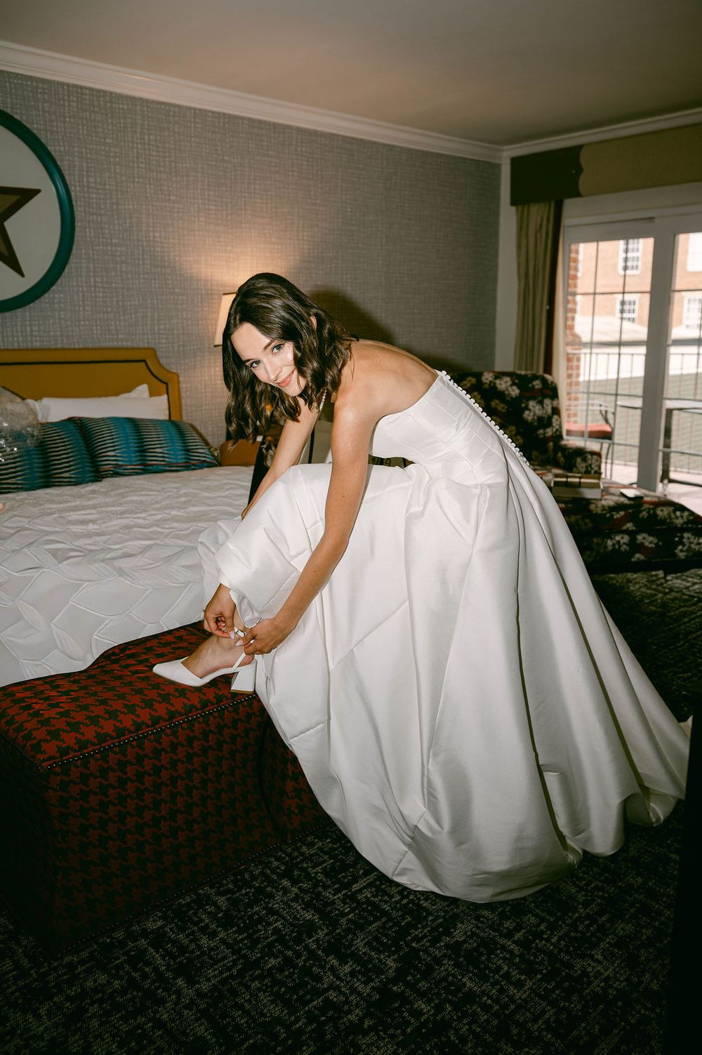 Bride in strapless white ball gown adjusting her shoe in an elegant hotel room before her wedding ceremony.