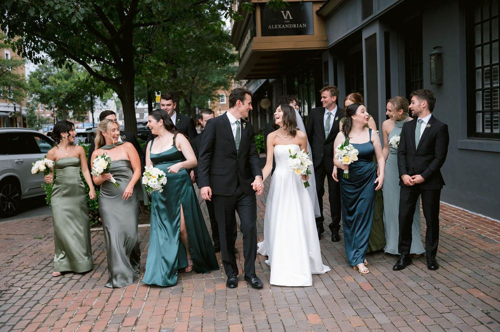 Wedding party walking on brick sidewalk, bridesmaids in green gowns, groomsmen in suits, bride in white dress.