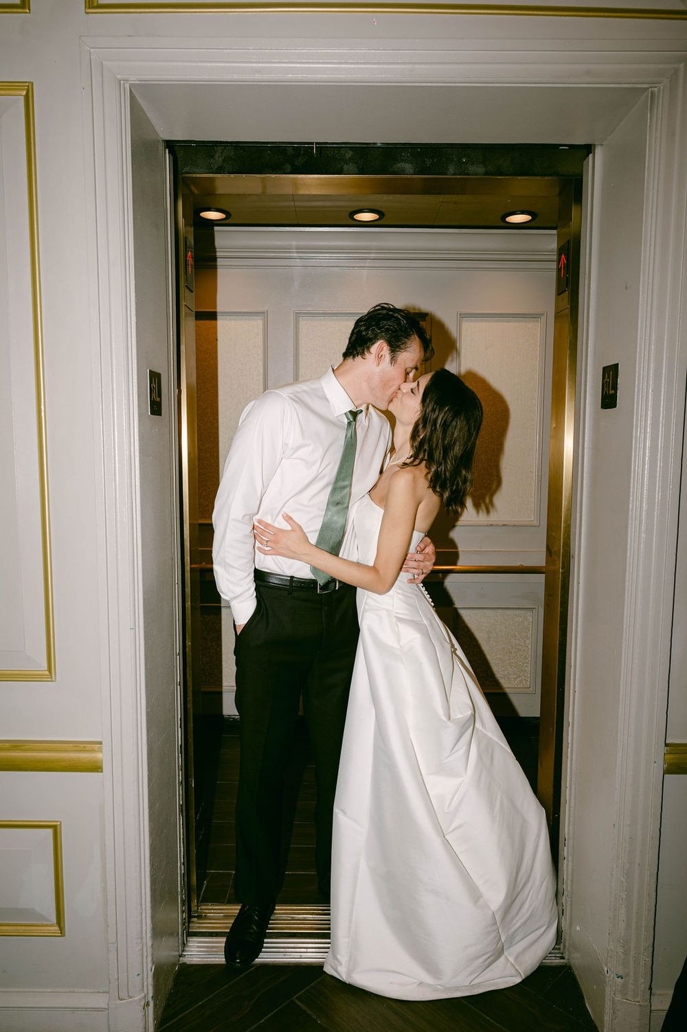 Newlywed couple sharing a romantic kiss in a gold elevator, bride in white gown, groom in dark suit with green tie.