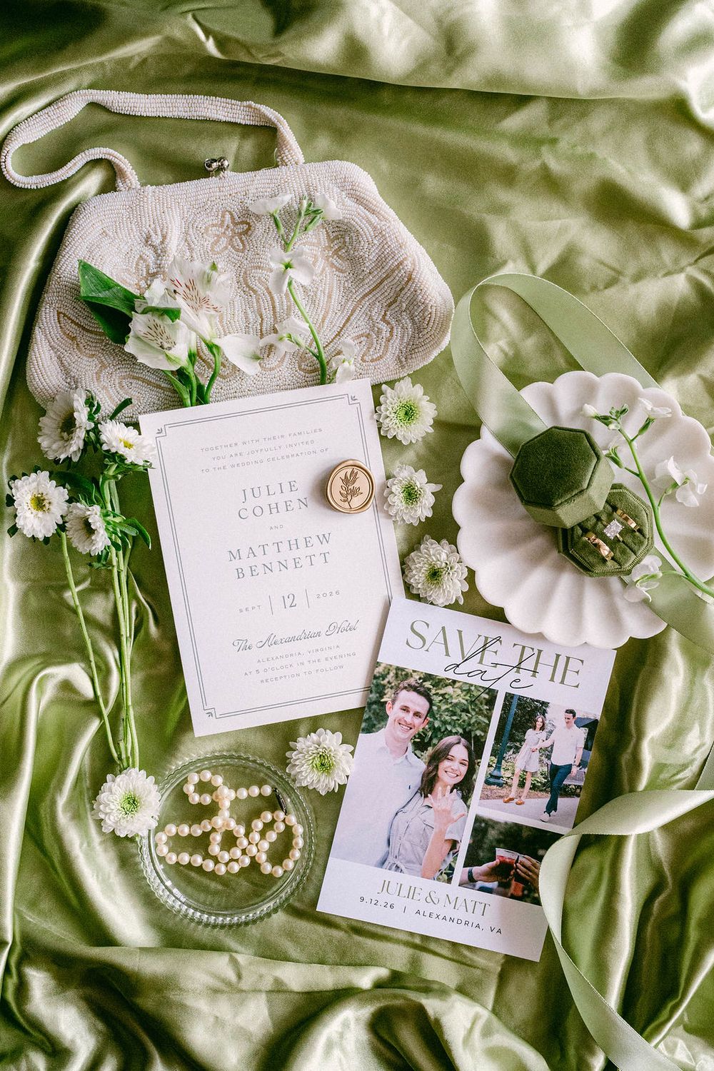 Wedding flat lay with invitation suite, save the date, pearl jewelry, floral clutch, and ring box on green silk.