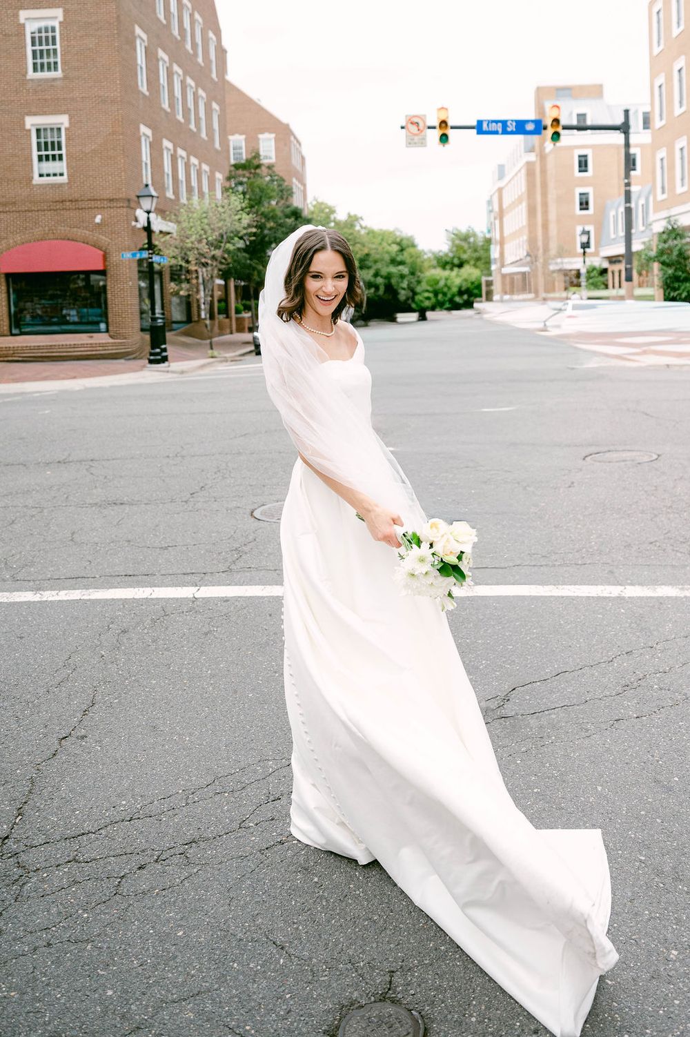 Bride in white gown and veil holds bouquet while crossing a city street intersection on her wedding day.