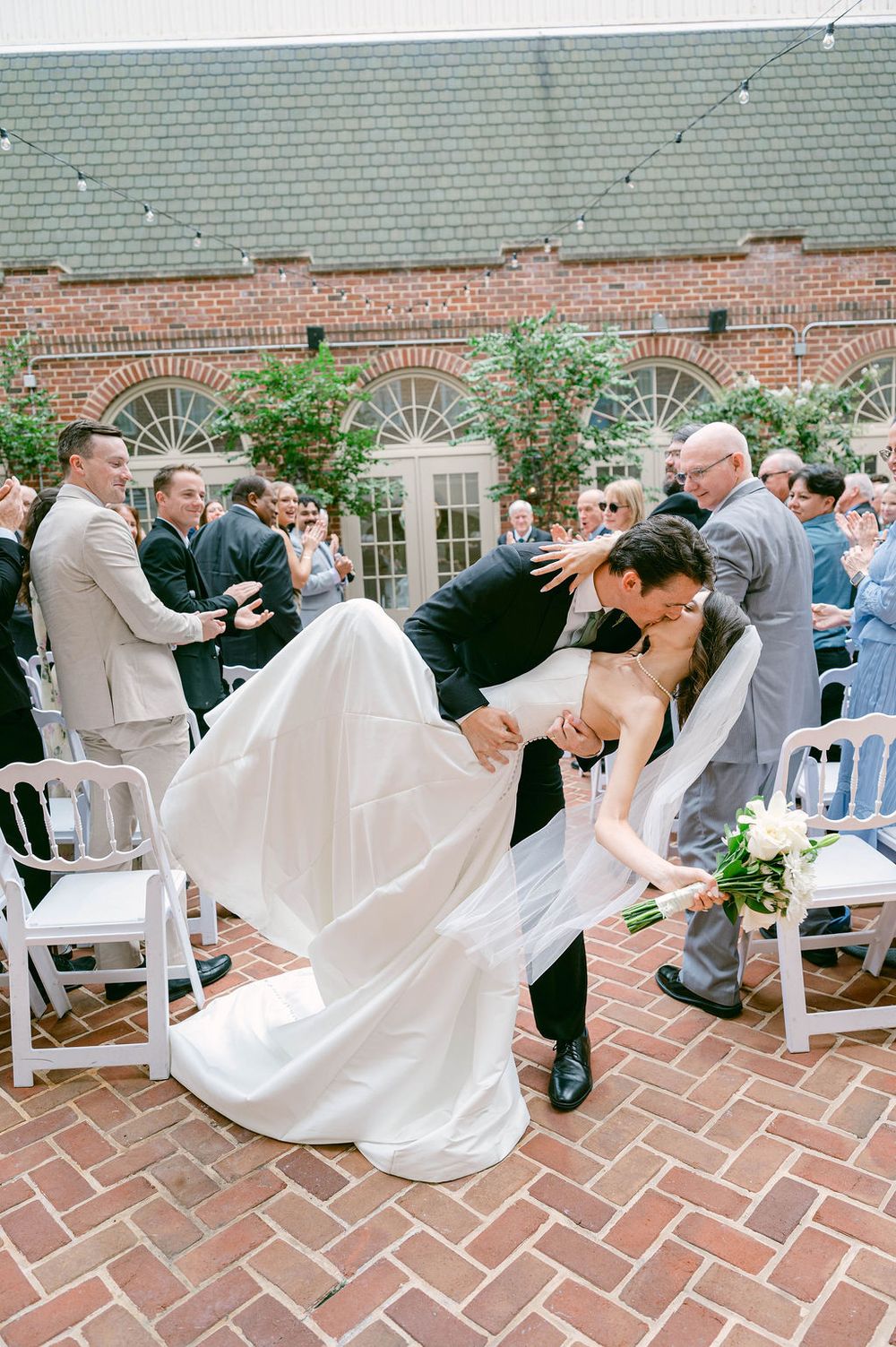 Groom dips bride for a romantic kiss during outdoor wedding ceremony on brick courtyard surrounded by guests.