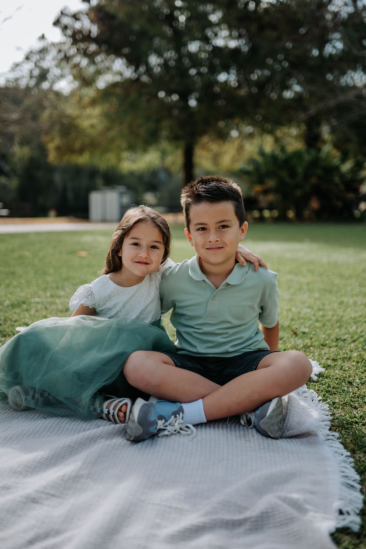 Two young siblings sitting together on a blanket in a park, smiling for a portrait photo outdoors.