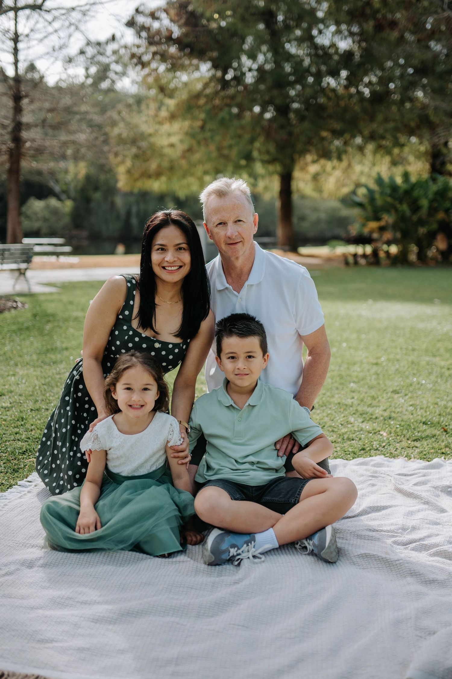 Family of four posing on a blanket in a lush green park, smiling warmly at the camera.