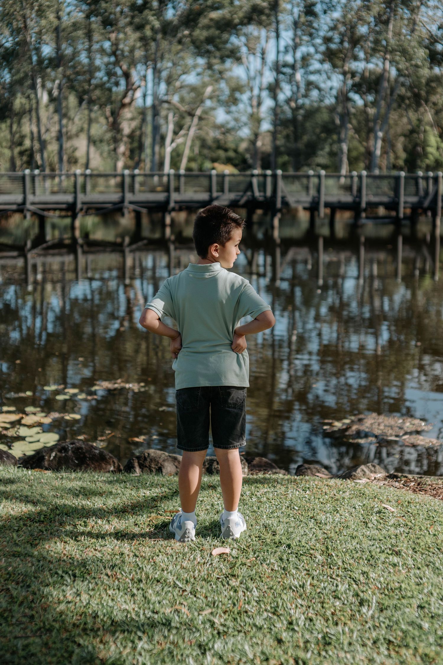Young boy in a sage green polo stands by a calm reflective lake in a sunny park.