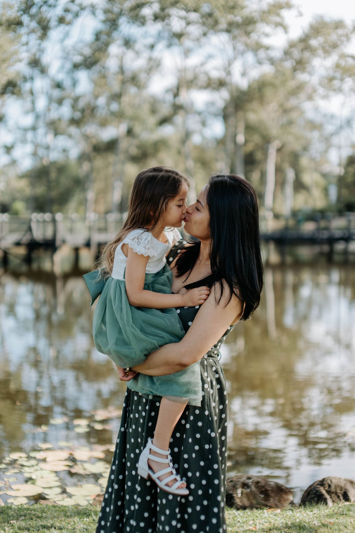 A mother holds and kisses her young daughter in a green dress by a serene park pond.