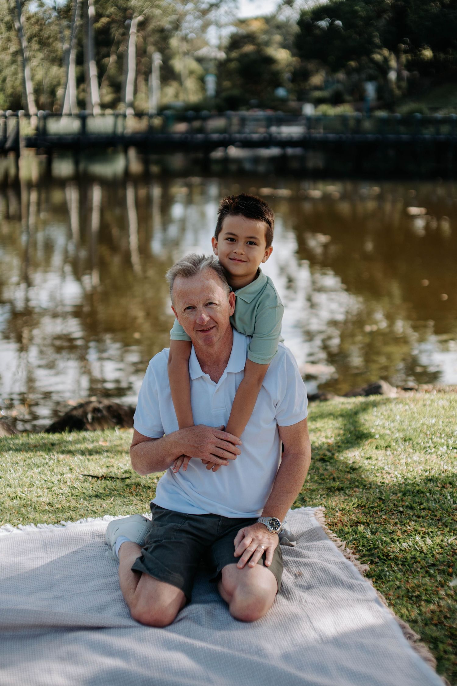 A smiling grandfather sits on a blanket by a river while his young grandson hugs him from behind in a park.