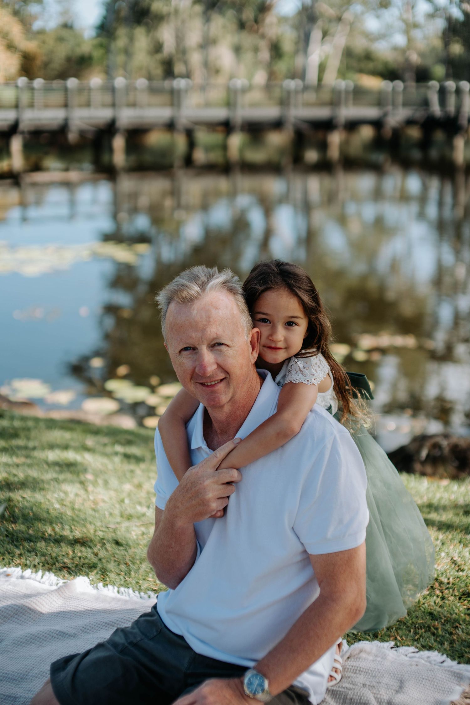 Man in white polo shirt smiling with young girl hugging him from behind near a serene pond with a wooden bridge.