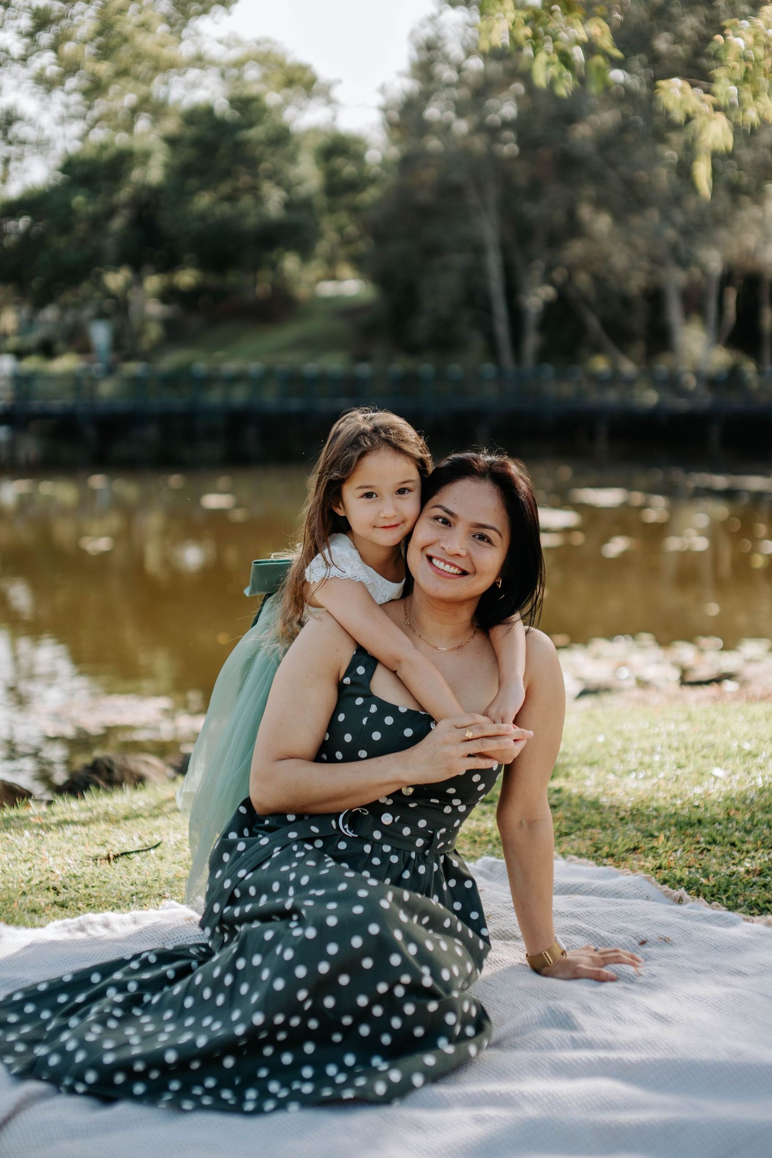 A woman in a polka dot dress sits on a blanket by a pond as a young girl hugs her from behind in a park.