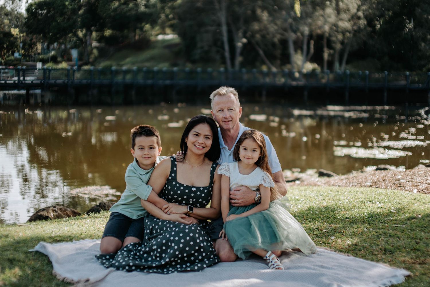 Family of four smiling together on a blanket by a serene pond in a lush green park setting.
