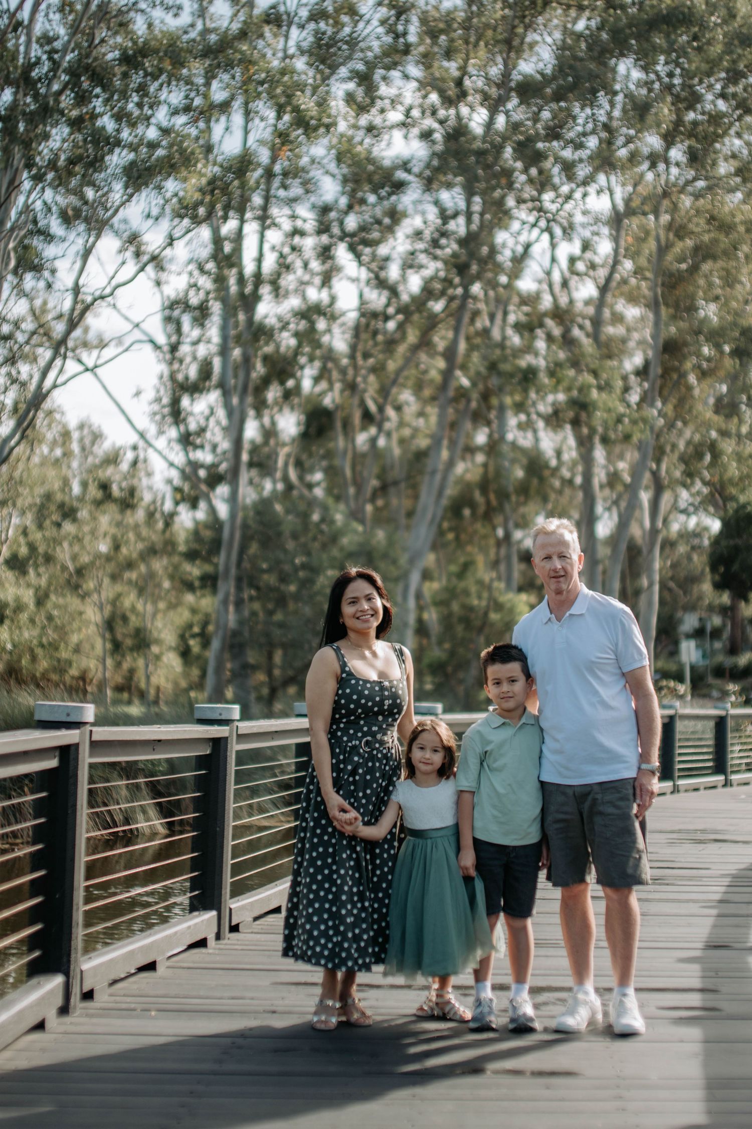 Happy family of four standing together on a wooden bridge surrounded by tall trees.