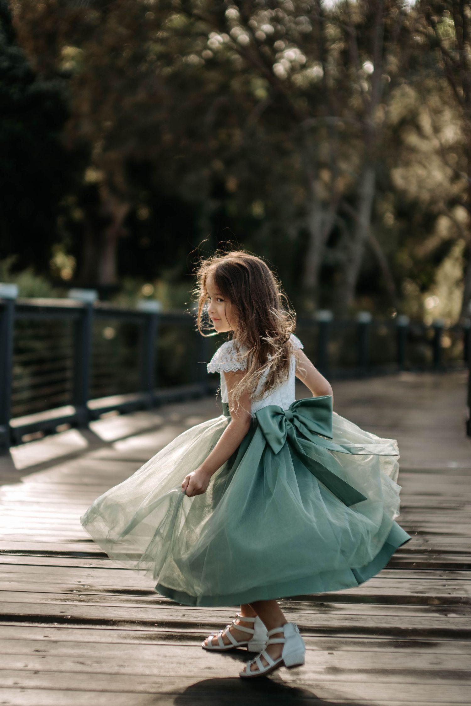 Young girl in white lace top and green tulle skirt with bow twirling on wooden bridge outdoors.