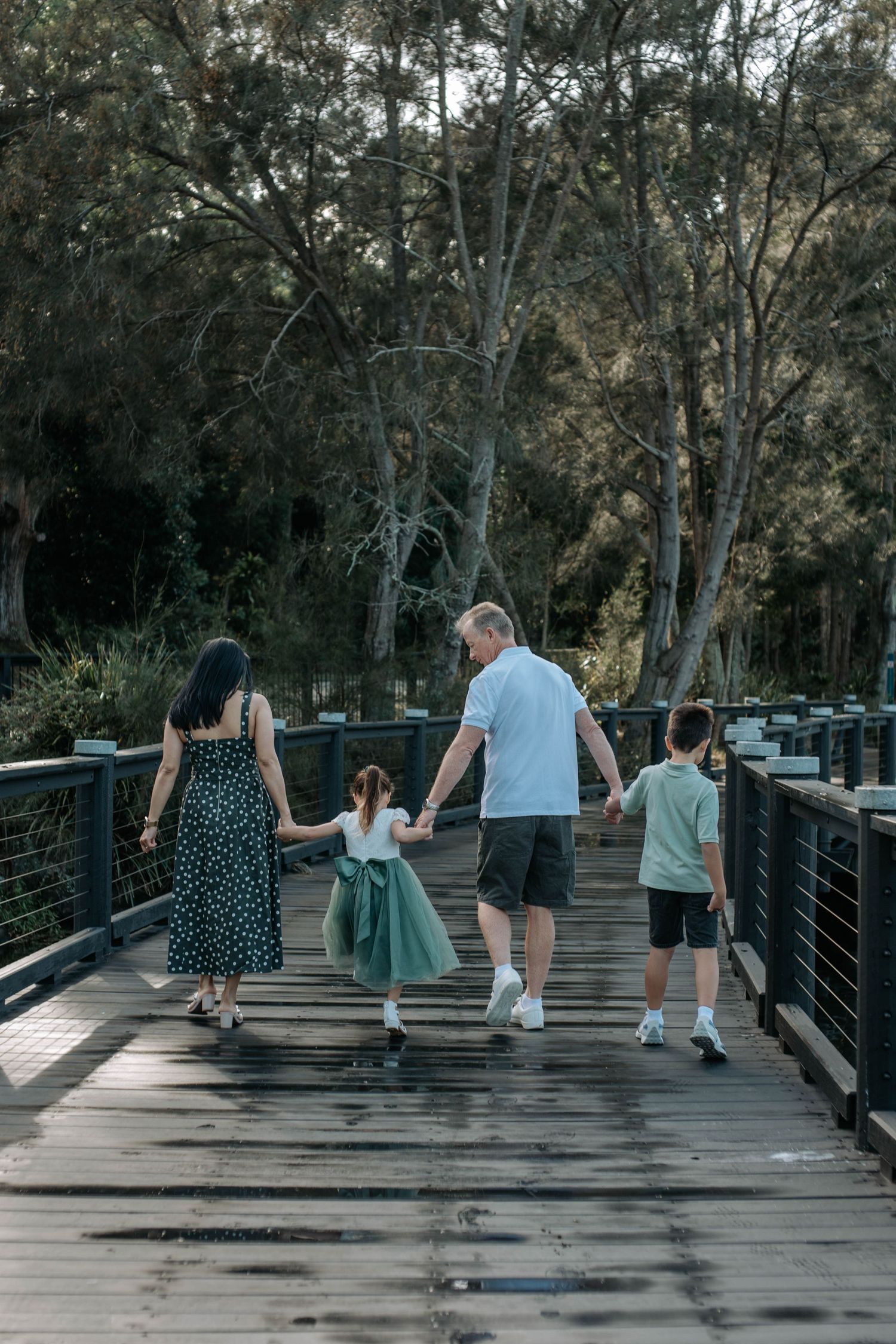 Family of four walking hand-in-hand across a wooden bridge surrounded by lush green trees.