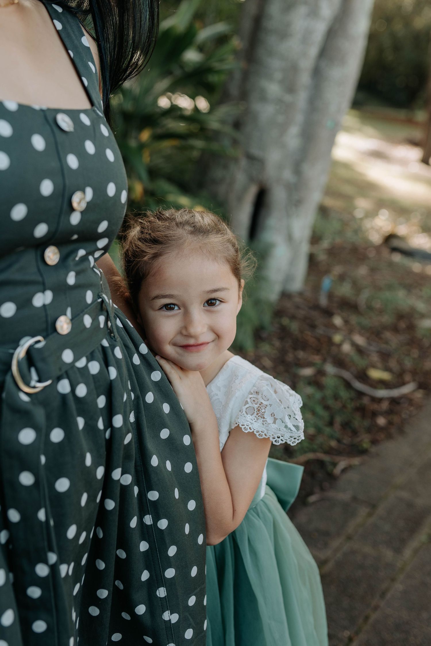 A cute young girl in a green tutu peeks out from behind her mothers green polka dot dress outdoors.