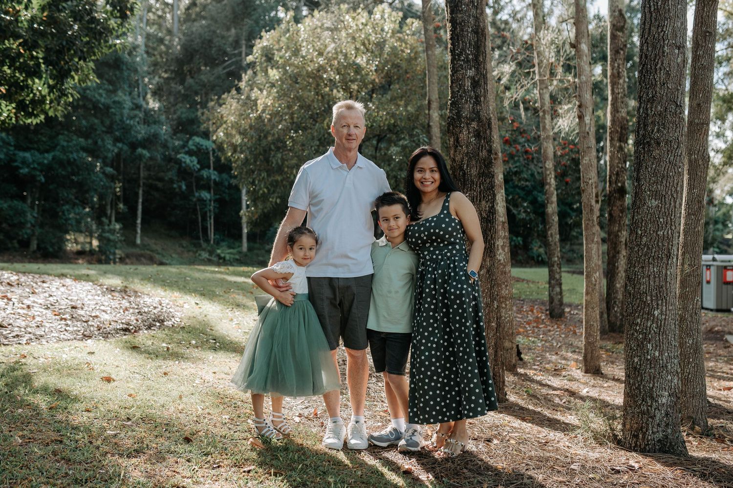 Family of four smiling together outdoors among tall trees in a lush green park setting.
