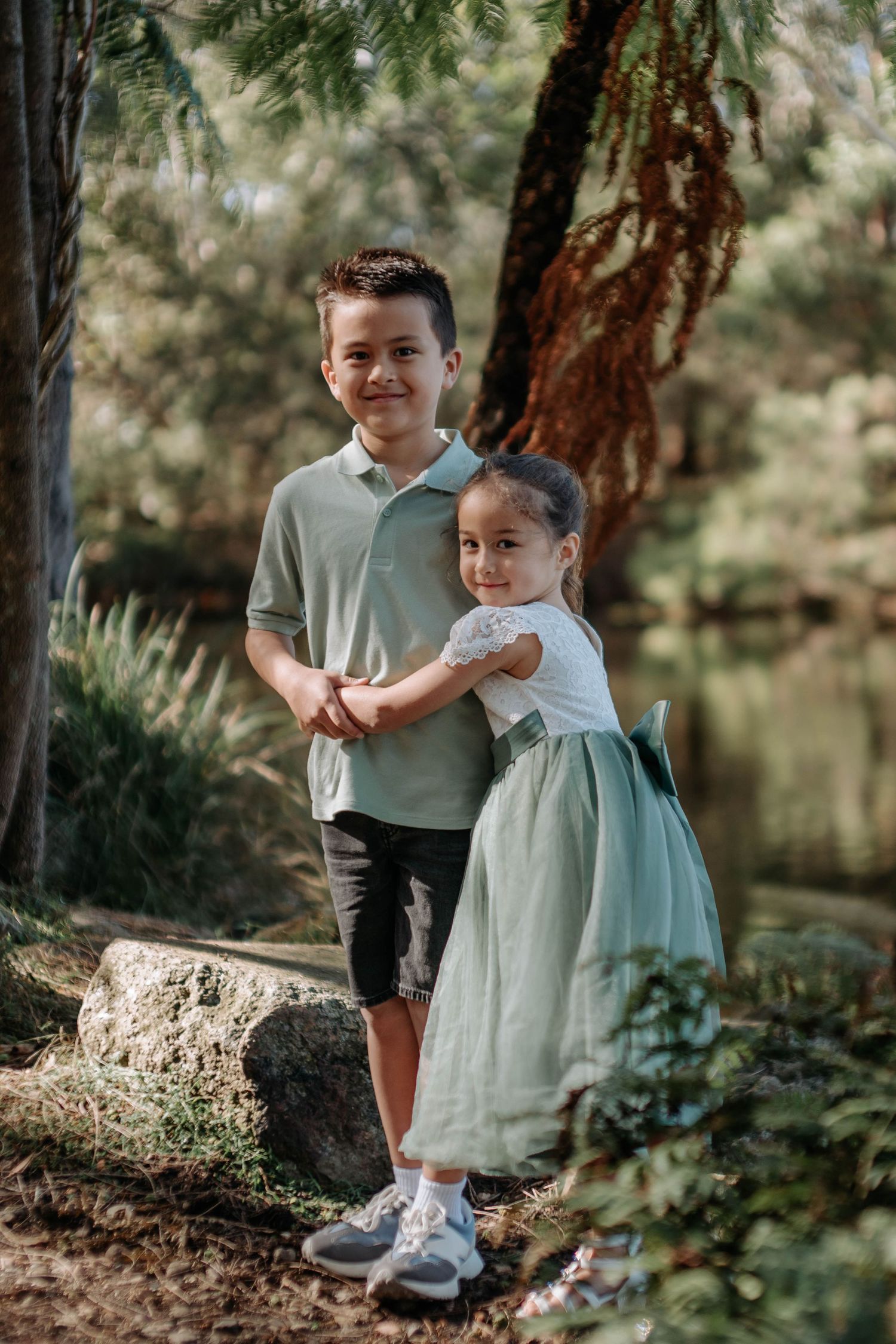 Two children, a boy in a sage polo and a girl in a mint dress, pose together in a lush garden park setting.