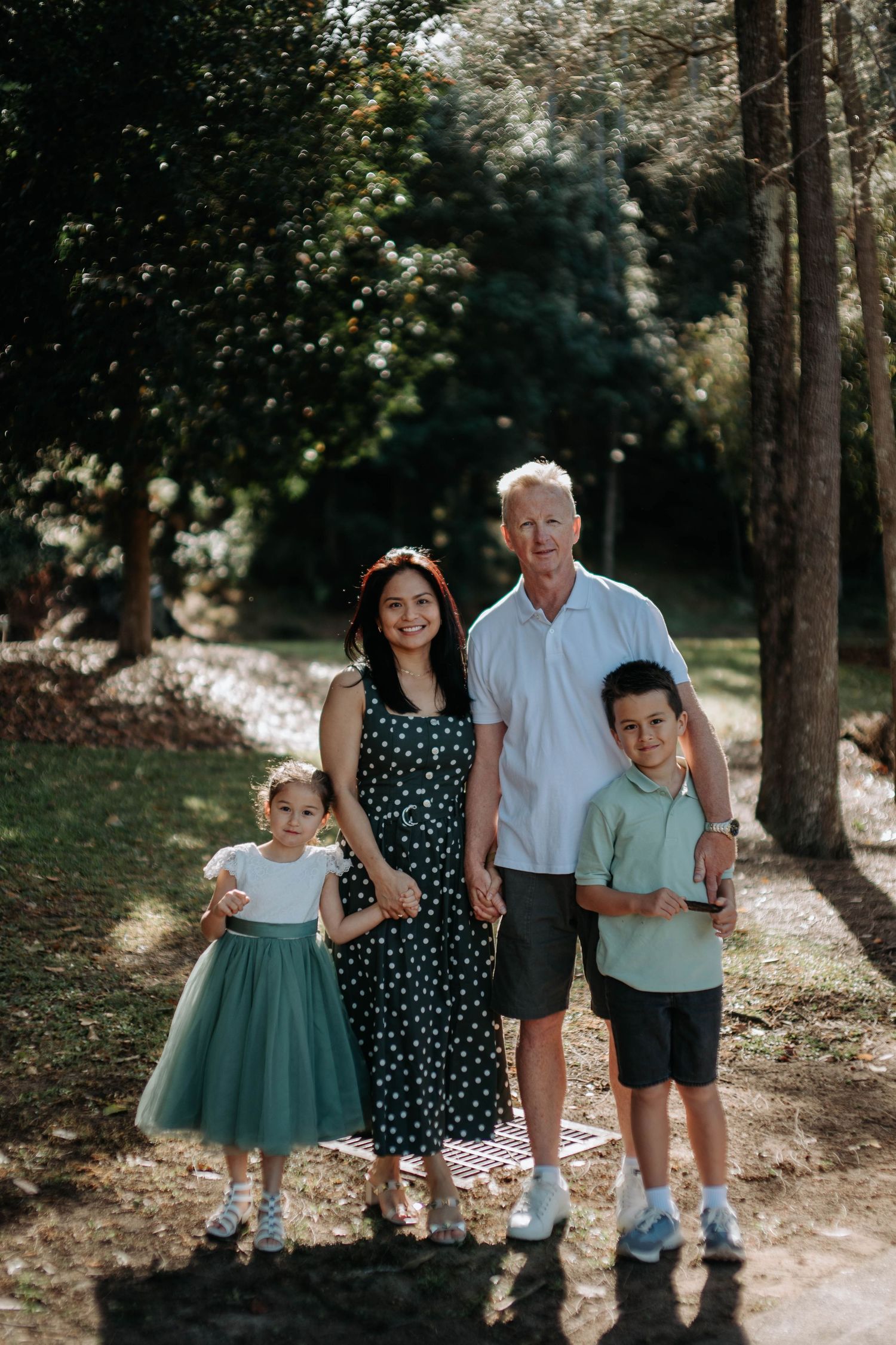 A happy family of four poses together on a sunny path surrounded by tall trees in a lush park.