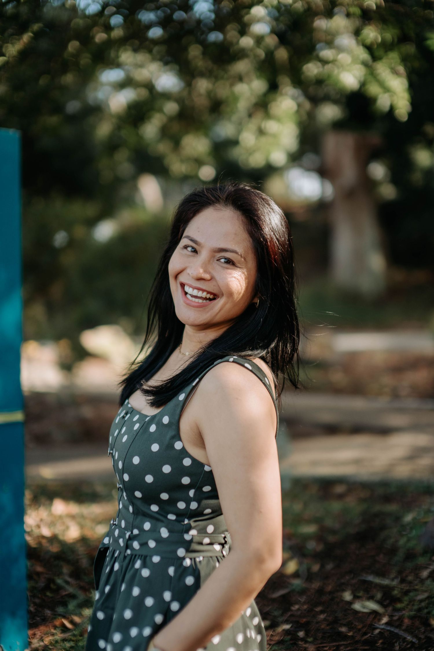 Smiling woman in polka dot dress standing outdoors among green trees in natural sunlight.