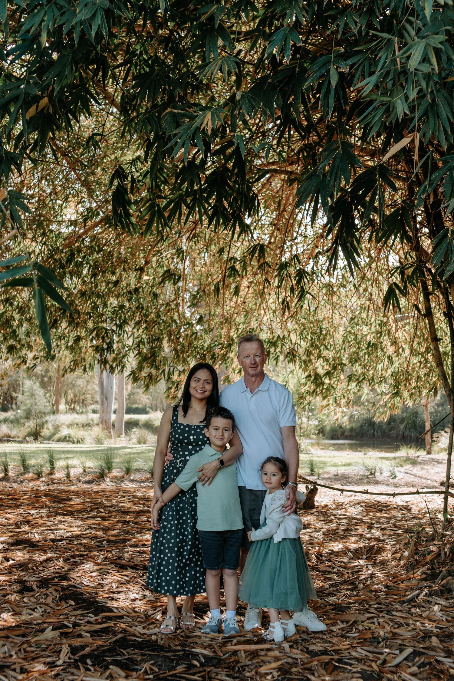 Family of four smiling under lush green tree canopy on fallen leaves in park setting.