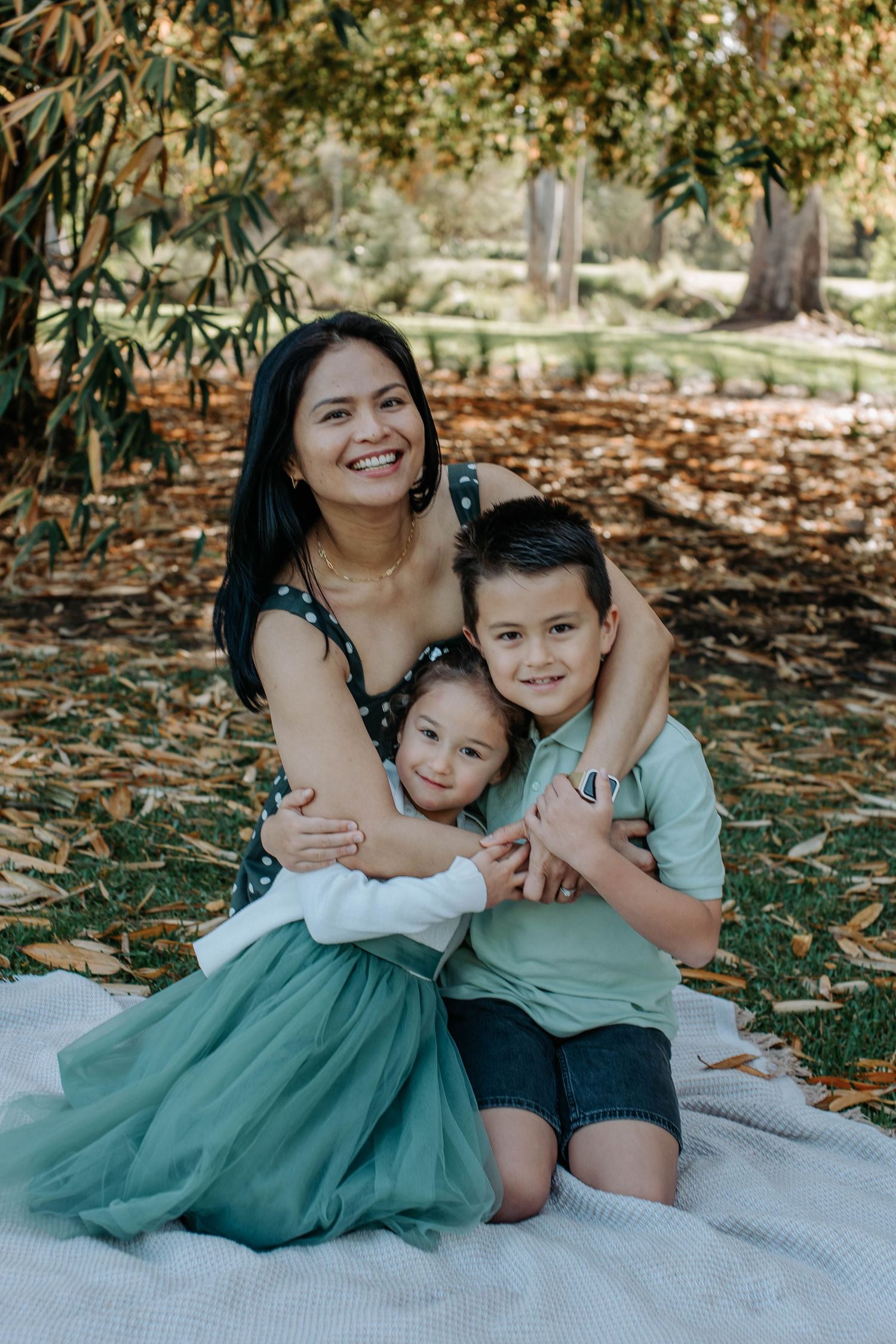 Smiling mother sits outdoors hugging her two young children under autumn foliage.