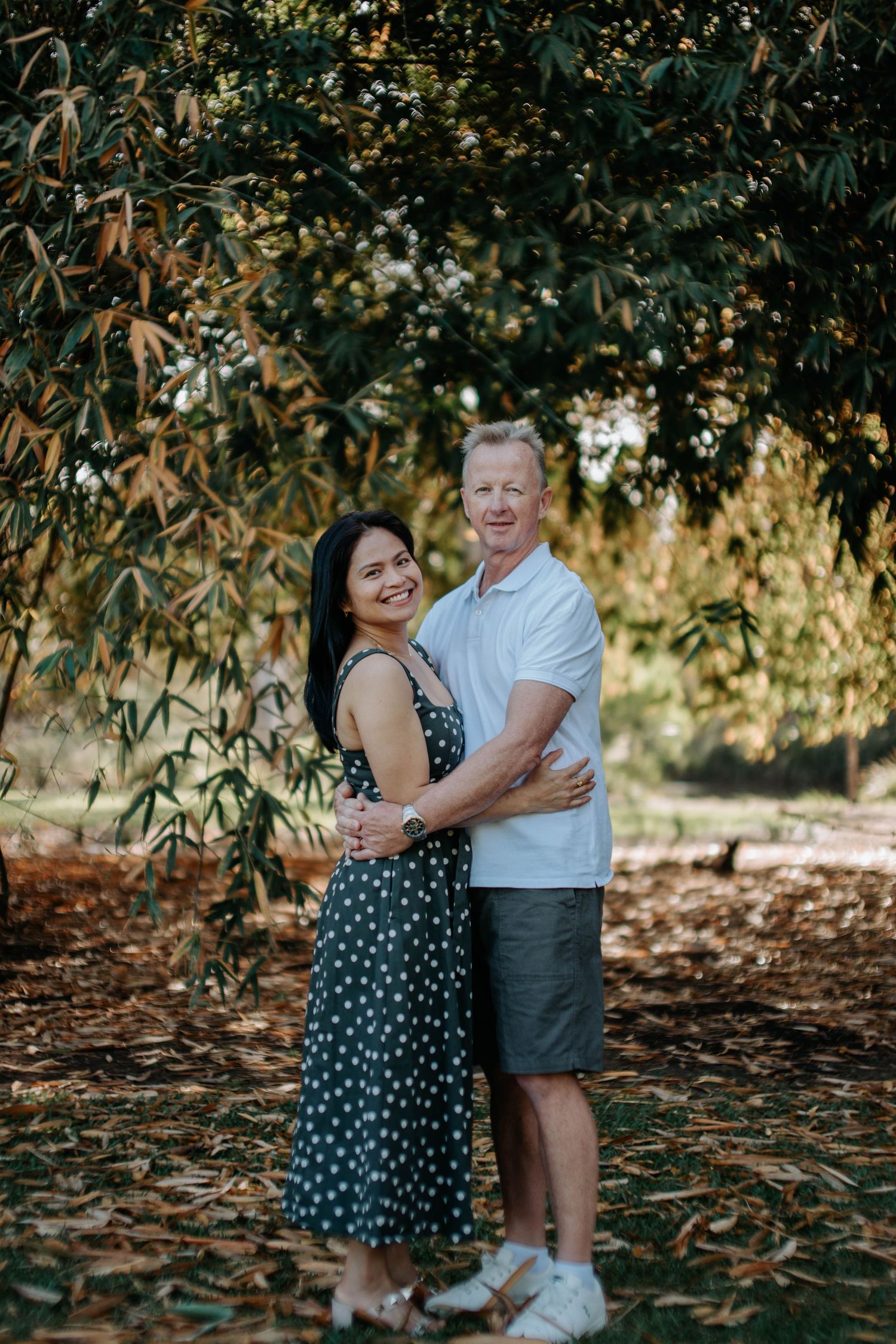 Couple embracing outdoors under lush green trees, woman in polka dot dress and man in white polo shirt.